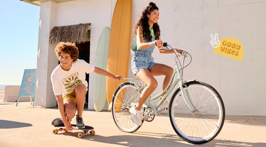 Two children, one on a bike and one on a skateboard, in front of a building with surfboards.