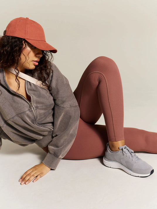 Person wearing a gray sweatshirt, maroon leggings, and a red cap sitting on a light-colored floor.