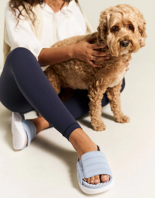 Person wearing blue slides and navy leggings sitting with a small brown dog on a light-colored floor.
