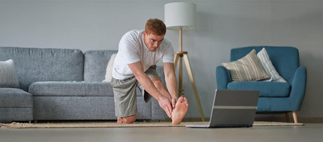 Man kneeling at home and reaching toward an extended bare foot while watching a laptop.