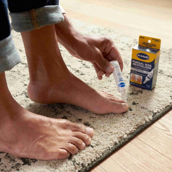 Person applying liquid from a small tube to a bare toenail while a box labeled Fungal Nail Revitalizer sits nearby on a rug