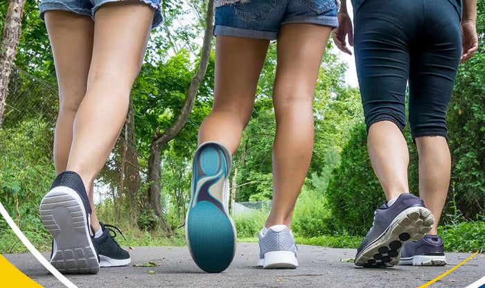 Three people walking along a paved path through a wooded park, viewed from behind, wearing shorts or leggings and athletic shoes.