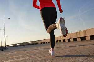 Woman runner in a red top and black leggings jogging across a bridge with one foot raised and long shadows stretching across the pavement.