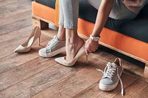 Person seated on an orange bench tying a beige sneaker, with another sneaker and a pair of beige high heeled shoes on the wooden floor nearby.